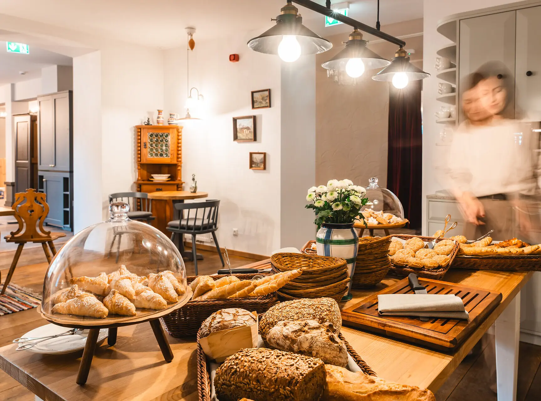 Cozy breakfast buffet with a variety of fresh bread and pastries displayed in wicker baskets, set in a rustic dining area with warm lighting and vintage decor.