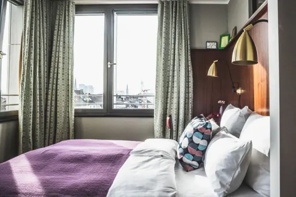 HENRI Düsseldorf Ein Bett mit Kissen und einer Lampe in einem Raum mit Fenstern.