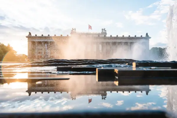 Das Alte Museum in Berlin bei Sonnenuntergang, gesehen durch die Wasserfontänen eines Springbrunnens mit Spiegelung im Vordergrund.