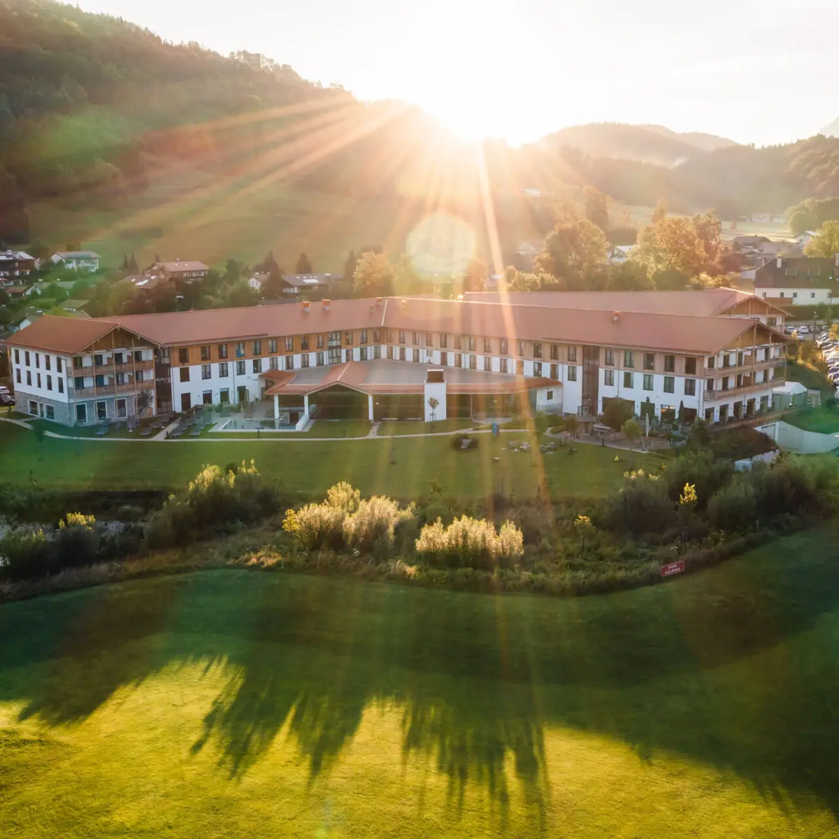 Luftaufnahme eines großen Hotels mit weitläufigem Garten in der Alpenlandschaft, Sonnenstrahlen strahlen über die Hügel und den Morgenhimmel