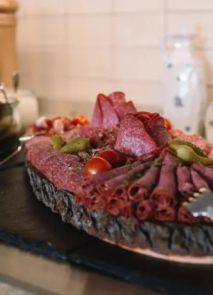Rustic wooden platter with a variety of sliced cured meats, presented on a wooden table alongside bread and cutlery.
