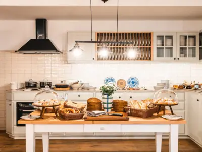 A beautifully designed kitchen with a rustic yet modern style, featuring white cabinetry, open shelves with cups, and a central wooden table displaying an assortment of fresh bread and pastries under glass covers.