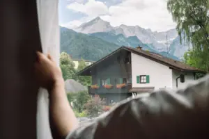 Ausblick im Salon Eine Hand hält einen Vorhang vor einem Fenster mit Bergen im Hintergrund.