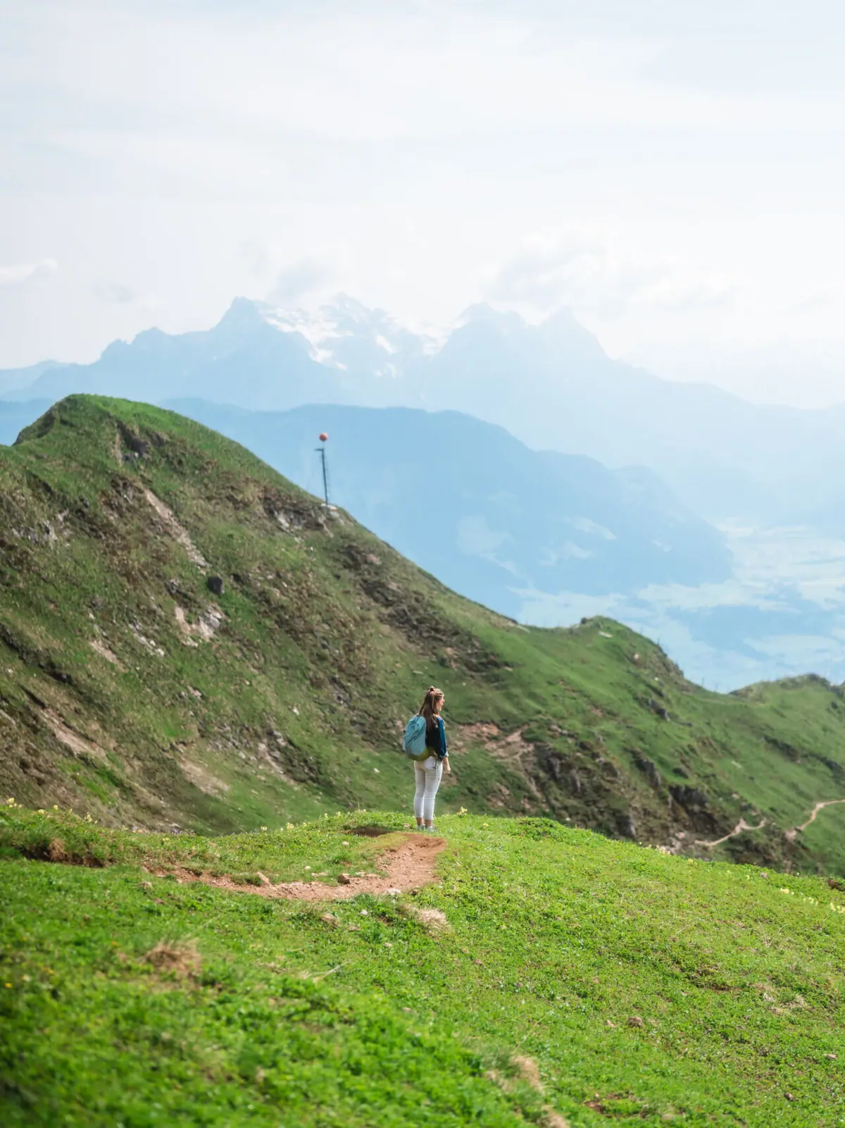 Eine Frau steht auf einem Pfad in einer grasbewachsenen Gegend mit Bergen im Hintergrund.