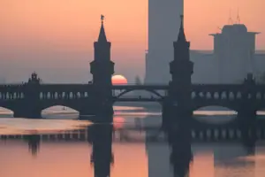 Silhouette der Oberbaumbrücke in Berlin bei Sonnenaufgang, wobei sich die Türme und die rötliche Sonne im ruhigen Wasser der Spree spiegeln.