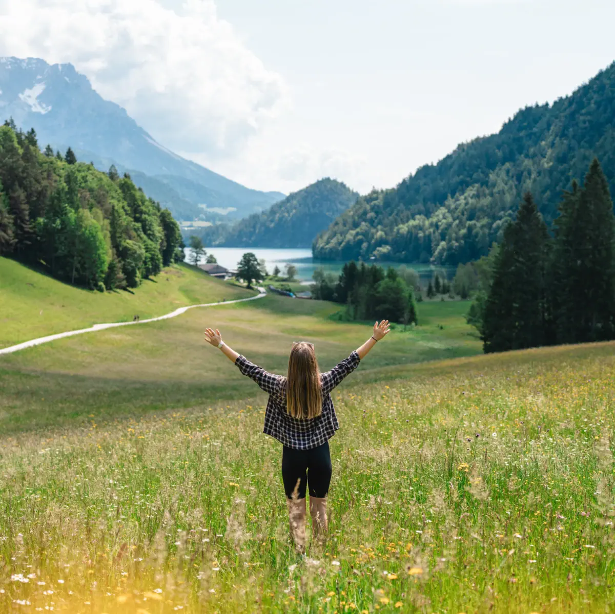 Eine Frau steht in einem Feld und streckt die Arme in die Höhe und im Hintergrund sind Berge zu sehen.