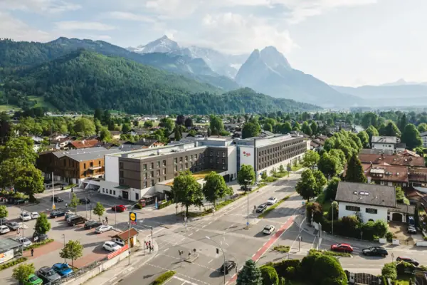 Das aja Garmisch-Partenkirchen mit vielen Gebäuden und Bäumen unter einem bewölkten Himmel sowie Bergen im Hintergrund.