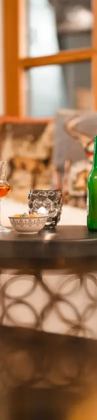 Cozy evening setup featuring a small round table with a glass of liqueur, a green beer bottle, a decorative candle holder, and a bowl of snacks.