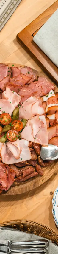 Rustic wooden platter with a variety of sliced cured meats, presented on a wooden table alongside bread and cutlery.