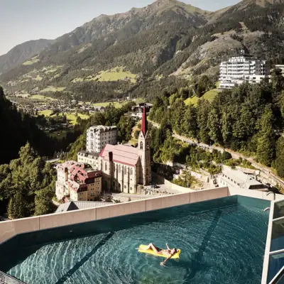 Eine Person schwimmt mit einer gelben Luftmatratze in einem Rooftop-Infinity-Pool auf einem hohen Gebäude mit Bergen im Hintergrund.