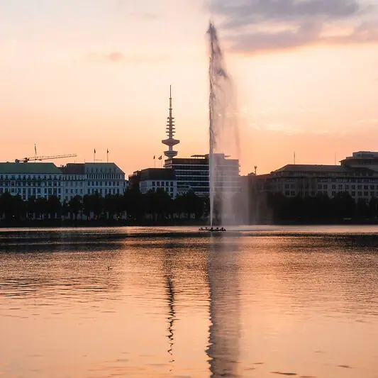 Springbrunnen in einem Gewässer mit Gebäuden im Hintergrund.