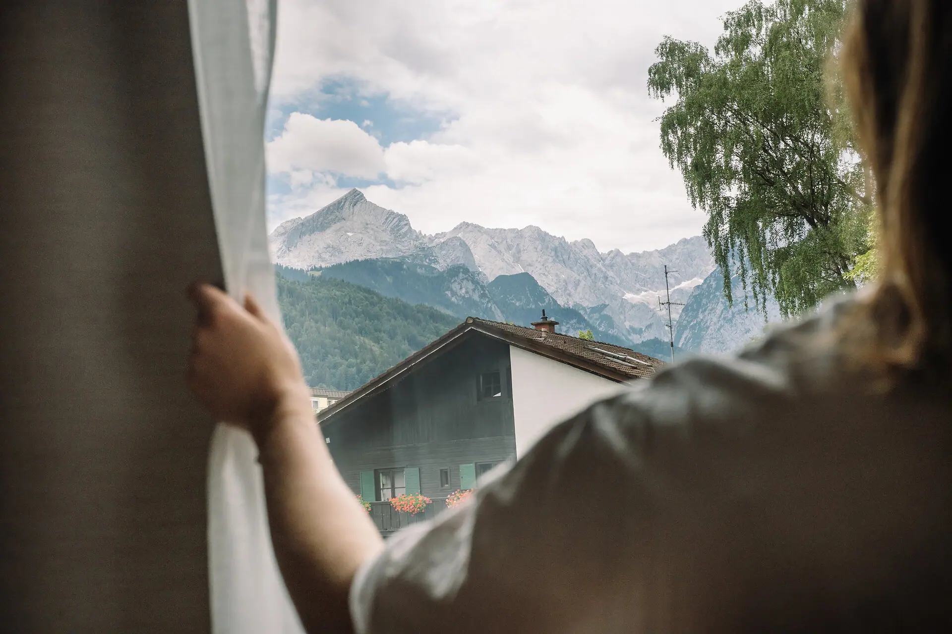 Eine Hand öffnet ein Fenster mit Bergpanorama im Hintergrund.