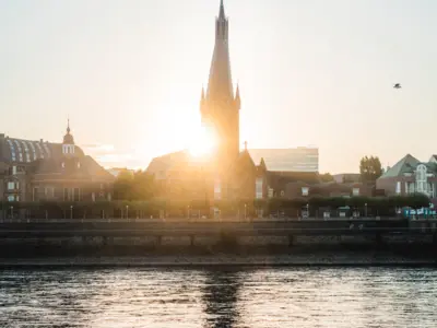 Rheinromantik und Großstadtflair – Düsseldorf weiß, wie man begeistert. Schnappen Sie sich unsere Black Deals und erleben Sie die Stadt von ihrer schönsten Seite.Schon ab 51 € p. P. inkl. Frühstück Stadtansicht mit hohem Turm und einem Gewässer bei Sonnenuntergang.