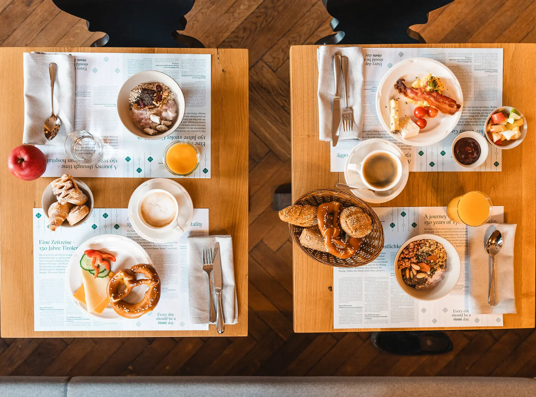 Top view of a breakfast table for two, featuring a variety of dishes including pretzels, fresh rolls, cheese, cold cuts, muesli, fresh fruit, and croissants, accompanied by coffee, orange juice, and water. 