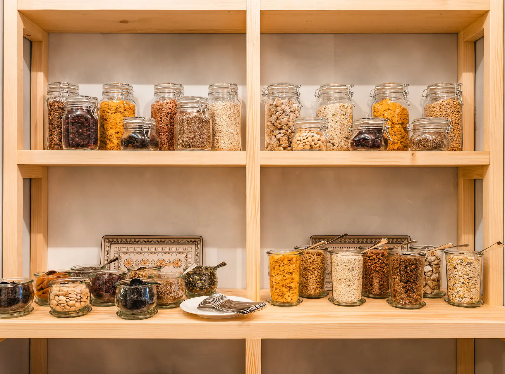 Breakfast station with neatly arranged jars of cereals, nuts, dried fruits, and toppings on wooden shelves.