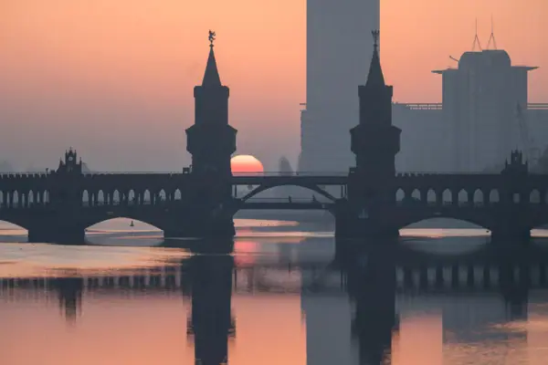 Silhouette der Oberbaumbrücke in Berlin bei Sonnenaufgang, wobei sich die Türme und die rötliche Sonne im ruhigen Wasser der Spree spiegeln.