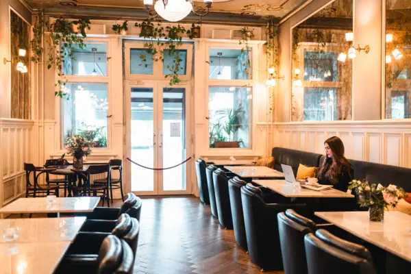 Eine junge Frau arbeitet am Laptop in der stilvollen Hotellobby des HENRI Berlin, umgeben von klassischen Holztäfelungen, Marmortischen und hängenden Grünpflanzen im Belle-Époque-Stil.