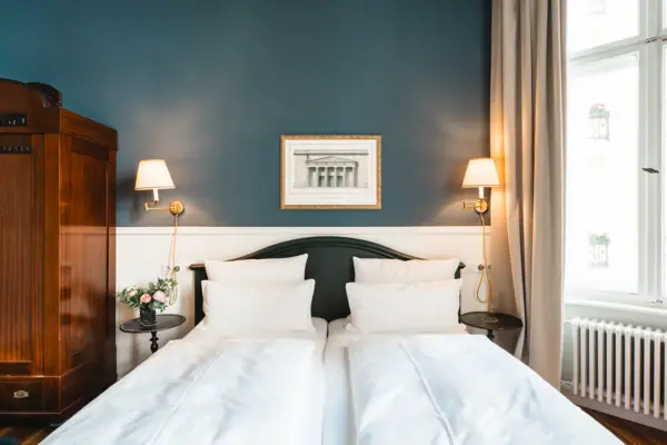 Frontale Ansicht eines stilvollen Doppelbetts im HENRI Berlin mit tiefblauer Wand, klassischem Holzschrank, symmetrischen Wandleuchten und einem gerahmten Architektur-Druck.