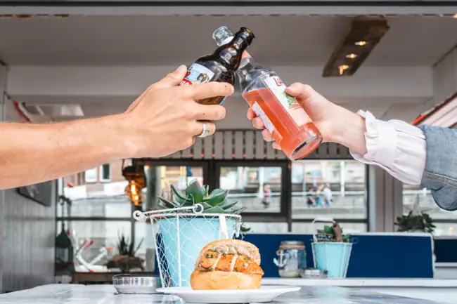 Fischbrötchen Hamburg Zwei Hände stoßen mit Flaschen an einem Tisch an.