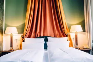 HENRI Hotel Berlin Zimmer Detail Baldachin Detailansicht eines herrschaftlichen Hotelbetts im HENRI Berlin mit einem prachtvollen orange-goldenen Baldachin vor einer grünen Wand und klassischen Nachttischleuchten.
