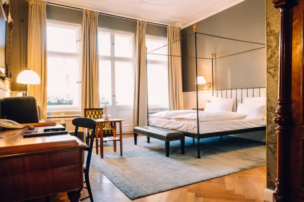 Großzügiges, lichtdurchflutetes Hotelzimmer im HENRI Berlin mit klassischem Himmelbett, Fischgrätparkett und stilvollen Vintage-Möbeln vor hohen Fenstern.