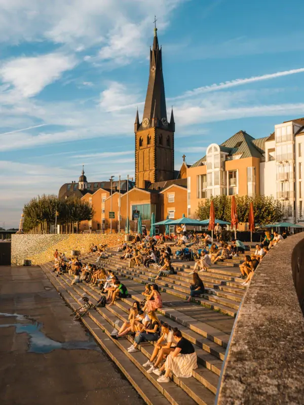 Rheinufer Düsseldorf Eine Gruppe von Menschen sitzt auf Treppen vor einem Gebäude.