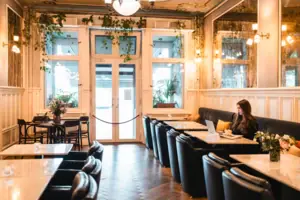 Eine junge Frau arbeitet am Laptop in der stilvollen Hotellobby des HENRI Berlin, umgeben von klassischen Holztäfelungen, Marmortischen und hängenden Grünpflanzen im Belle-Époque-Stil.