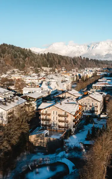 A picturesque winter village nestled in a valley surrounded by snow-covered mountains. Charming alpine-style buildings with snow-dusted roofs are scattered amidst trees, with majestic peaks rising in the background under a bright blue sky.