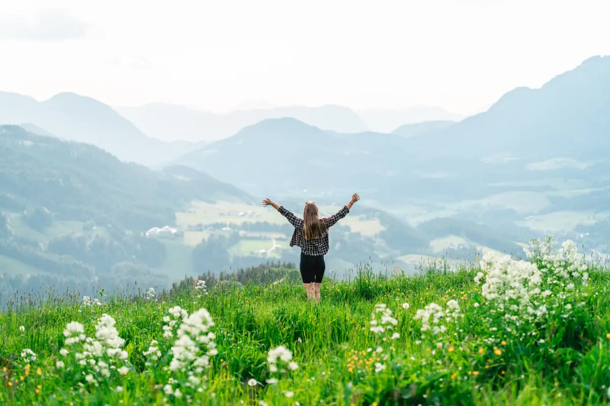 Eine Frau steht auf einer bergwiese und hebt die Arme während Sie auf das umliegende Bergpanorama schaut.