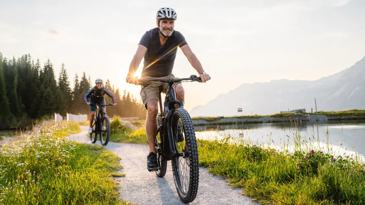 Ein Mann fährt auf einem Weg Fahrrad, im Hintergrund sind ein See und Berge zu sehen.