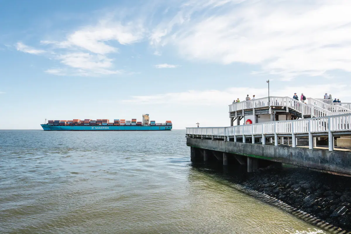 Ein Containerfrachtschiff fährt auf dem Wasser vor einem Steg.